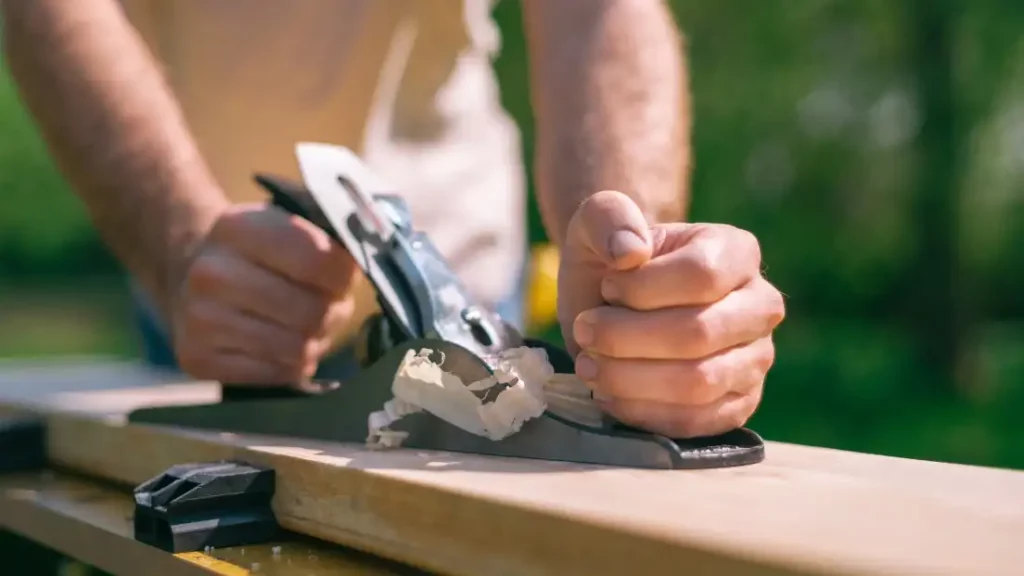 A man planning a piece of wood by hand with a jack plane with his hands and wood shavings in focus