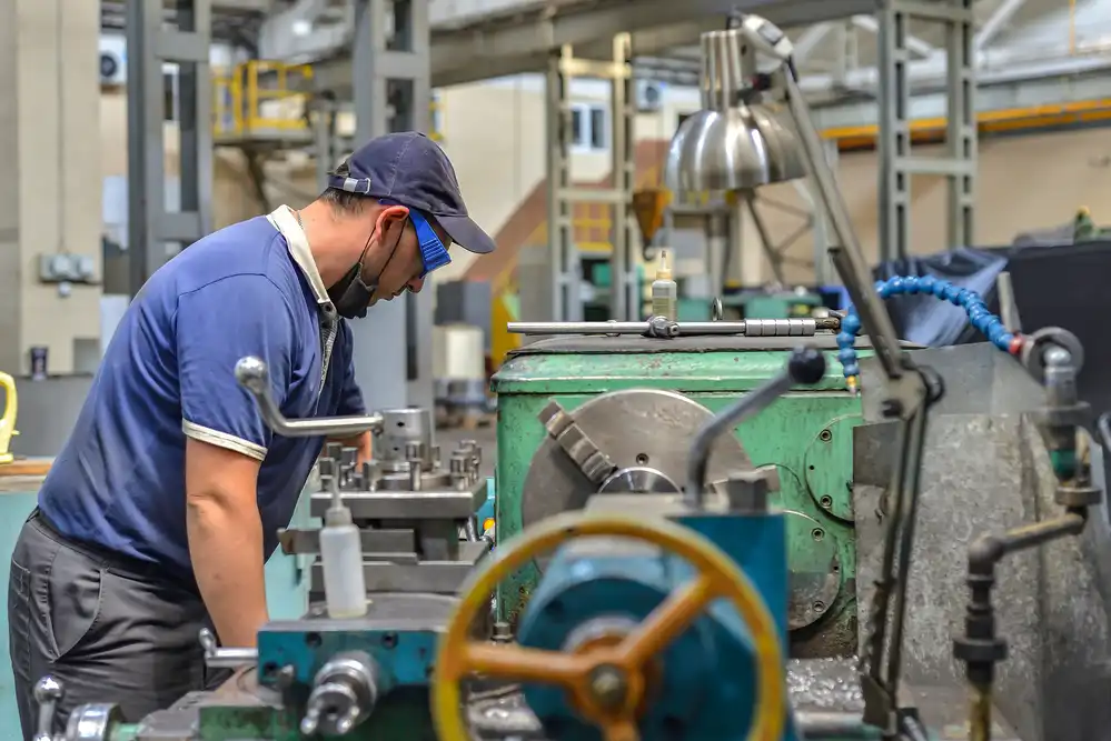 Working turner wearing a protective antiviral mask on a lathe in a workshop. The machine operator at the workplace controls the equipment